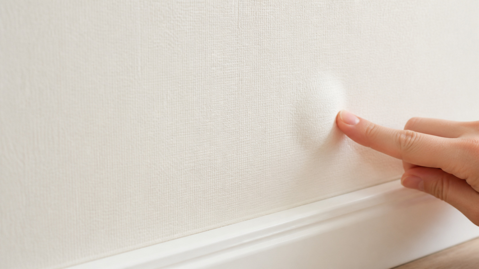 Hand deutet auf eine kleine Wölbung in einer intakten hellen Tapetenfläche oberhalb der Fußleiste